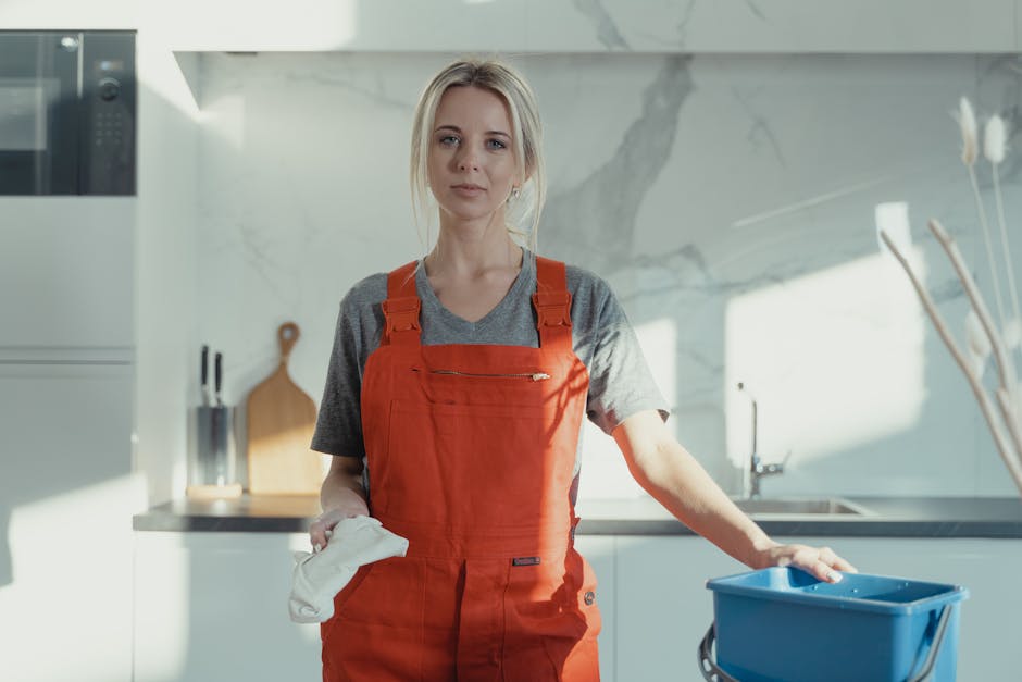 A professional cleaner in a modern kitchen, standing confidently with a neutral expression, wearing a red apron over a gray t-shirt. She holds a white cleaning cloth in her right hand and rests her left hand on a blue mop bucket. The kitchen features a white marble backsplash with subtle gray veining, stainless steel appliances, including a built-in microwave, and a wooden cutting board leaning against the wall. The surfaces are clean and polished, illuminated by natural light from a nearby window, highlighting the pristine condition of the countertops and stainless steel sink, emphasizing hygiene and surface cleaning as part of domestic or commercial cleaning services provided by Carpet Cleaners W9.