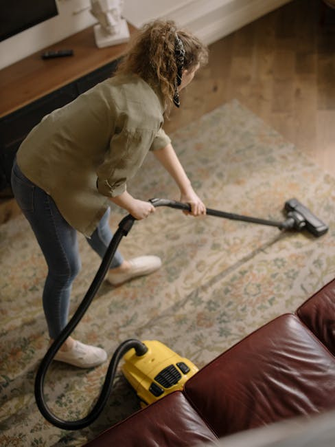 A woman with curly hair, wearing a beige jacket, blue jeans, and white shoes, is performing surface cleaning using a yellow vacuum cleaner with a hose attachment on a patterned area rug in a living room. The room has wooden flooring adjacent to the rug, a brown leather sofa partially visible in the foreground, and a wooden entertainment unit against the wall behind her. The space appears well-lit with natural light, and the carpet is clean with no visible dust or stains. The image demonstrates domestic cleaning practices suitable for the services offered by Carpet Cleaners W9 for deep cleaning and sanitisation of residential surfaces.