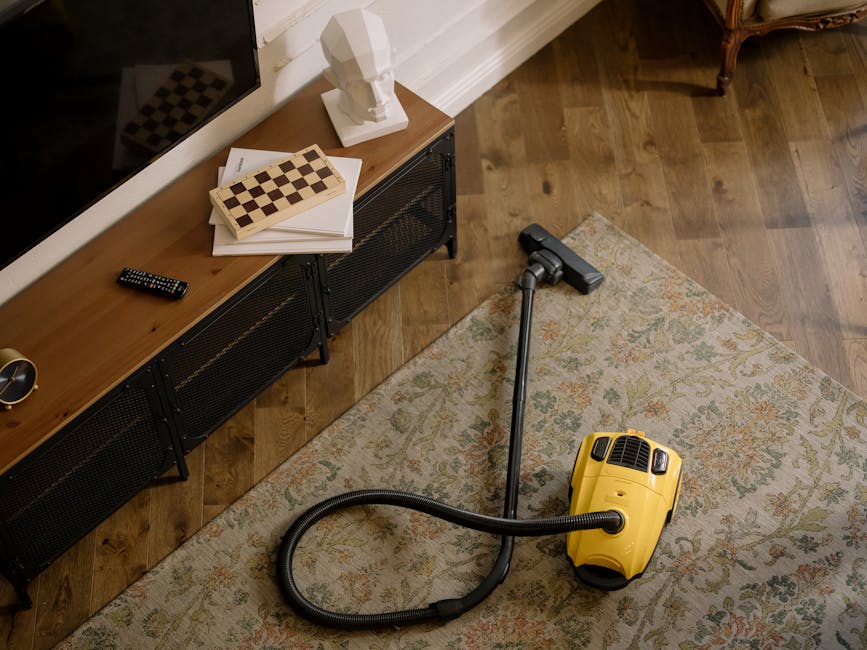 A yellow vacuum cleaner with a hose attachment is positioned on a patterned area rug over a wooden floor in a living room. To the left, a low wooden sideboard with black metal legs holds a white bust sculpture, a television remote control, and a chessboard. The room features a white baseboard along the wall, which is partly visible. The area appears clean and well-maintained, with natural lighting illuminating the space. This setting demonstrates surface cleaning and maintenance typical of domestic cleaning services provided by Carpet Cleaners W9, situated near Warwick Avenue station in Maida Vale W9.