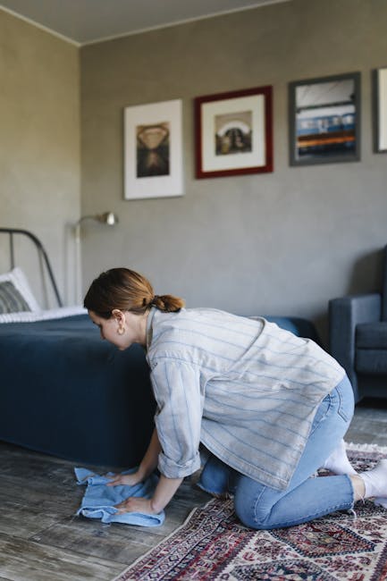 A woman with light brown hair tied back, wearing a light blue and white striped shirt and jeans, is kneeling on a patterned area rug while using a blue cloth to clean a dark wooden floor in a residential living room. The room features a grey wall decorated with three framed pictures and a beige ceiling. To her right, there is a dark blue upholstered sofa and an armchair, with a window visible in the background allowing natural light into the space. The scene reflects domestic cleaning activities focused on floor surface cleaning and maintaining hygiene, consistent with services provided by Carpet Cleaners W9 for problem resolution in Maida Vale flats, W9.