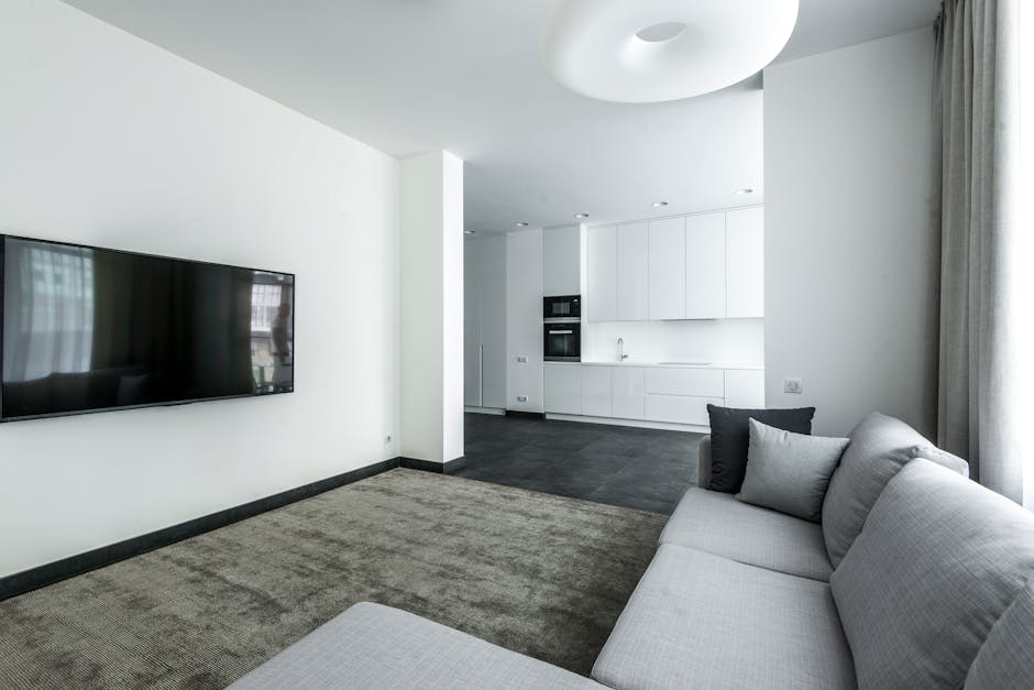 A modern, minimalist living room with a light grey fabric sofa adorned with dark grey and light grey cushions, sitting on a textured beige carpet. Across from the sofa, a large flat-screen television is mounted on a white wall. Adjacent to the living area is an open-plan kitchen with white cabinetry, built-in oven, and a stainless steel sink, all set against a dark tiled floor. Natural light filters through a large window dressed with light grey curtains, illuminating the space evenly. The room appears clean and well-maintained, showcasing a setting suitable for surface cleaning, deep cleaning, and domestic sanitisation services offered by Carpet Cleaners W9 in Maida Vale flats, W9.