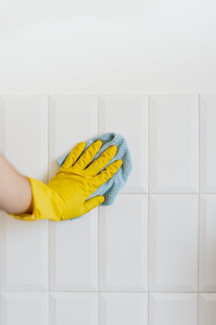 A close-up view of a person's hand wearing a bright yellow rubber glove, using a blue microfiber cloth to clean white ceramic wall tiles in a residential kitchen or bathroom. The wall tiles are arranged in a grid pattern, with visible grout lines, and appear freshly wiped, showing a shiny, spotless surface. The background features a plain white wall with minimal decoration, and the lighting is natural or overhead, highlighting the cleanliness and polishing process typical of surface cleaning services. This image exemplifies detailed domestic cleaning activities promoted by Carpet Cleaners W9, as part of their end of tenancy cleaning services in Sutherland Avenue Maida Vale, W9, emphasizing sanitation and maintenance of tiled surfaces.