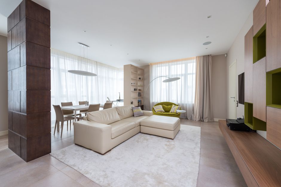 A spacious living room featuring a neutral beige carpet that appears clean and well-maintained, with sunlight streaming through a series of large bay windows adorned with striped pink and white curtai