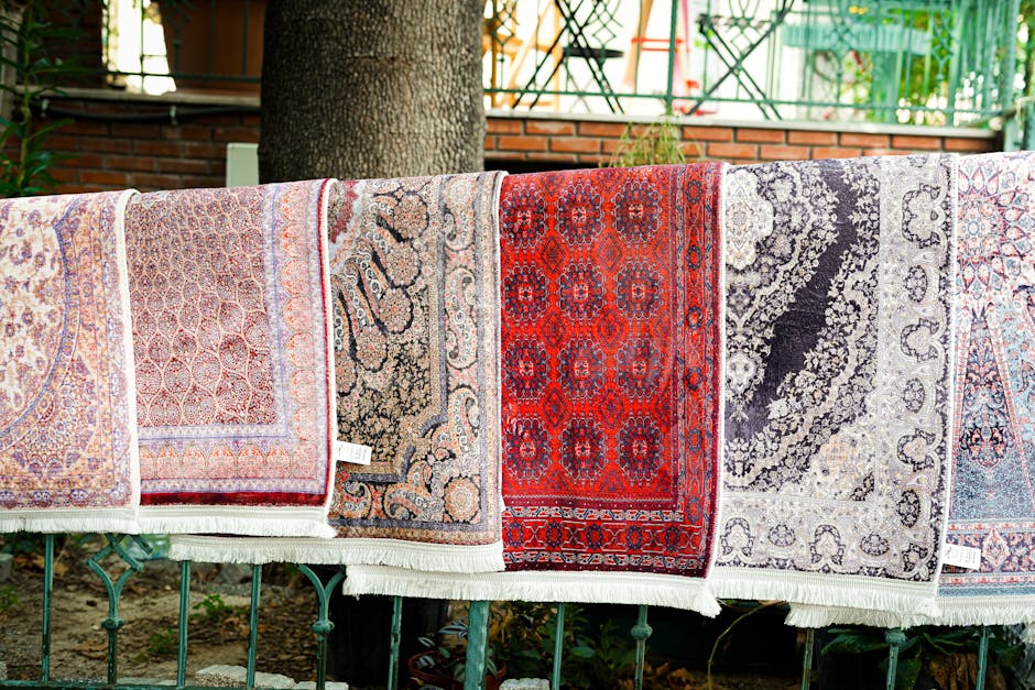 A row of colorful, patterned rugs hangs over a metal fence outdoors, showcasing intricate designs and vibrant colors including red, beige, black, and light blue. The rugs are drying in natural light, with some detail visible in the textures and patterns. In the background, there is a brick wall, green fencing, and foliage, indicating the laundry is set in a garden or backyard. The scene reflects clean, well-maintained textiles, demonstrating the type of surface cleaning and maintenance services offered by Carpet Cleaners W9 for domestic environments like Little Venice and Maida Vale, W9.