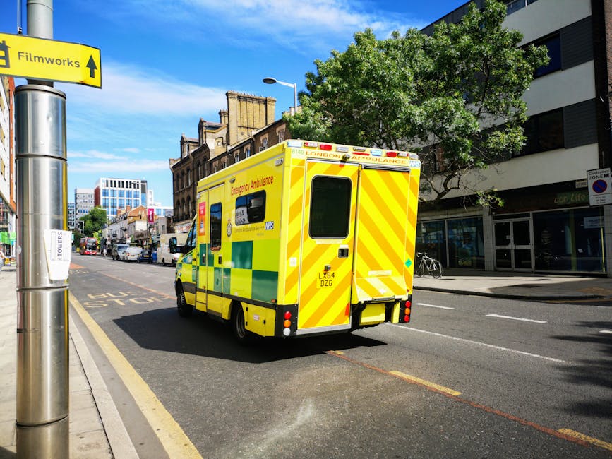 A bright yellow London emergency ambulance with green reflective markings is parked on the side of a busy urban street under clear blue skies. The ambulance is positioned near a bus stop with a yellow sign labeled 'Filmworks' pointing straight ahead. Surrounding the ambulance are modern multi-story buildings, including a white and black commercial building with glass windows on the right. There are trees with lush green foliage providing shade, and a few bicycles are parked along the sidewalk. The street is lined with vehicles, and the road surface appears clean and well-maintained, with visible lane markings. The scene is well-lit, capturing a typical day in a city environment, illustrating the importance of rapid response services like the ambulance in urban areas.