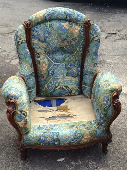An ornate armchair with a wooden frame featuring carved details, upholstered in a patterned fabric with shades of blue, green, and beige. The upholstery is heavily damaged, torn, and missing in several areas, revealing the internal padding and the frame beneath. The chair is situated outdoors on a paved surface, with a concrete background. The setting appears to be a street or driveway, and the lighting is natural. This image relates to domestic cleaning services offered by Carpet Cleaners W9, highlighting the importance of upholstery deep cleaning and maintenance.