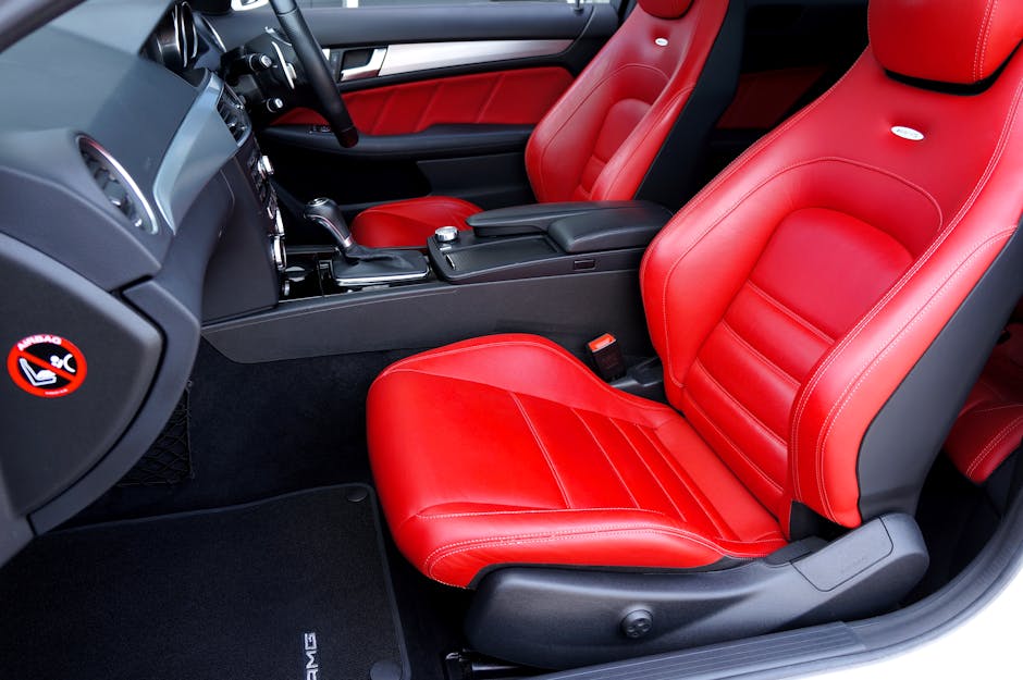 Close-up view of the front interior of a car featuring vibrant red leather seats with visible stitching, a black dashboard with control buttons, a gear shift, and a steering wheel. The seats appear clean and well-maintained, with no visible dust or stains. The black floor mats and door panels complement the red upholstery, and sunlight streams into the vehicle, highlighting the polished surfaces. This image demonstrates interior surface cleaning and maintenance services provided by Carpet Cleaners W9, specializing in deep cleaning and sanitisation of domestic and commercial vehicle interiors within the Elgin Avenue Maida Vale, W9 area.