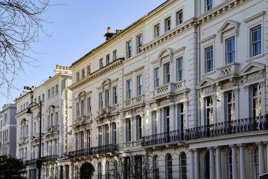Exterior view of a row of elegant Victorian-style residential buildings with white facades and ornate architectural details, including decorative moldings, balustrades, and columns, under clear blue sky. The buildings feature large, evenly spaced windows, some with small balconies with black wrought iron railings. The street is illuminated by vintage-style black lampposts, and leafless trees are visible in the foreground. The scene appears clean and well-maintained, reflecting a tidy urban residential environment, with sunlight casting soft shadows on the building surfaces. Carpet Cleaners W9 provides surface cleaning and deep cleaning services, ensuring high standards of hygiene and cleanliness consistent with Westminster Council rules for commercial cleaning in Maida Vale, W9.