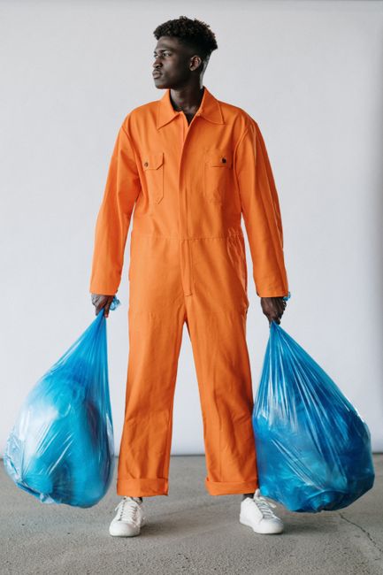 A person wearing an orange protective suit, black mask, white gloves, and sneakers is standing outdoors on a concrete pavement next to a gray brick wall, holding two large green trash bags filled with
